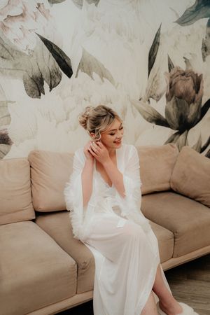 A bride in a flowing white dress sits elegantly on a tan couch during wedding preparations.