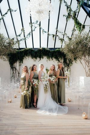 Bridal party poses under glass ceiling conservatory decorated with hanging greenery and lanterns.