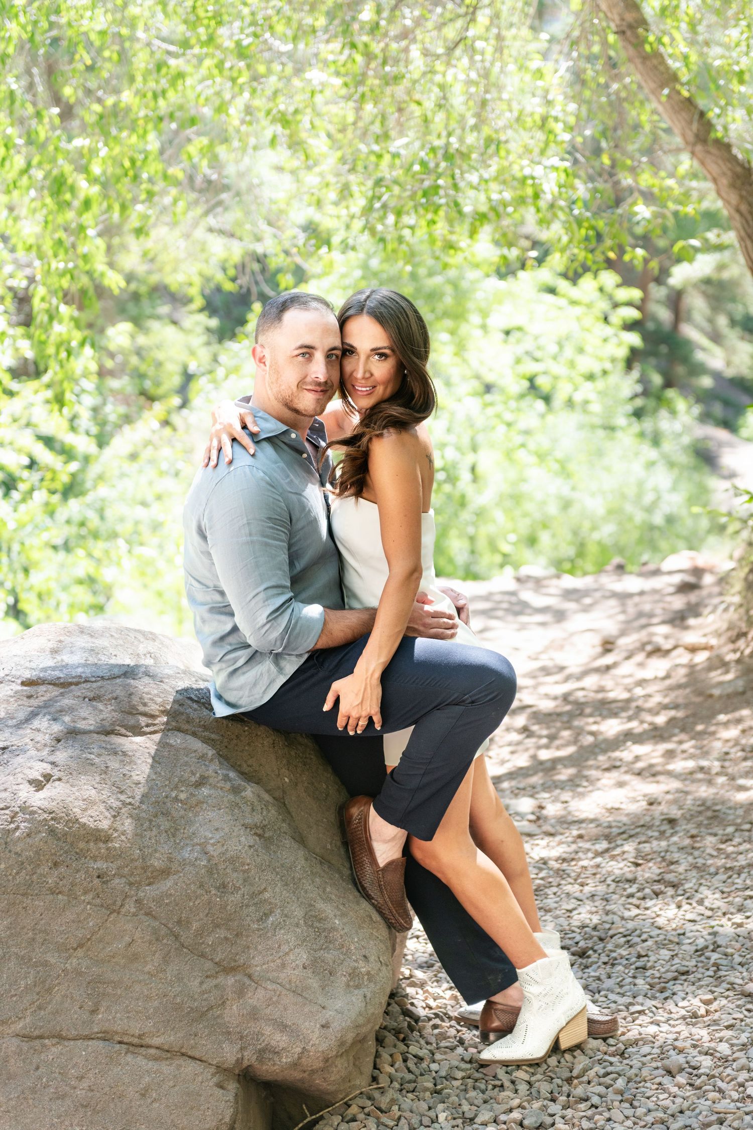 An eloping couple poses on a rock in a lush green park, the woman leaning on the the man's chest with her arms him and both are smiling.