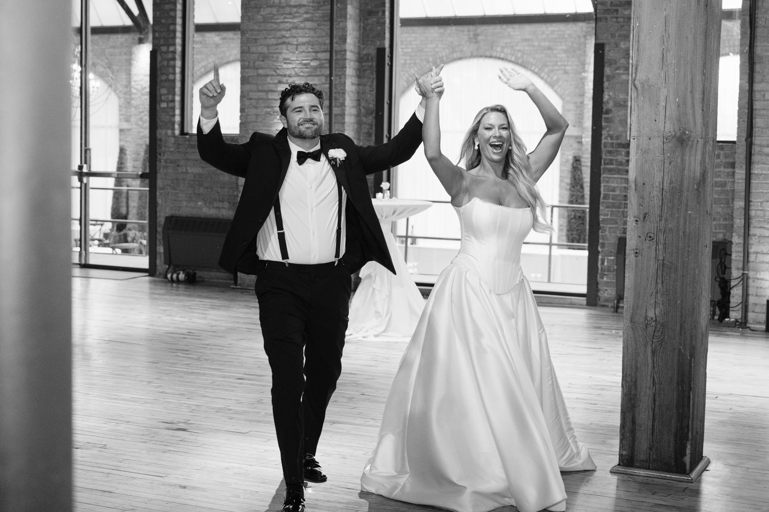 A wedding party celebrates in a rustic industrial venue with exposed brick walls and large windows.