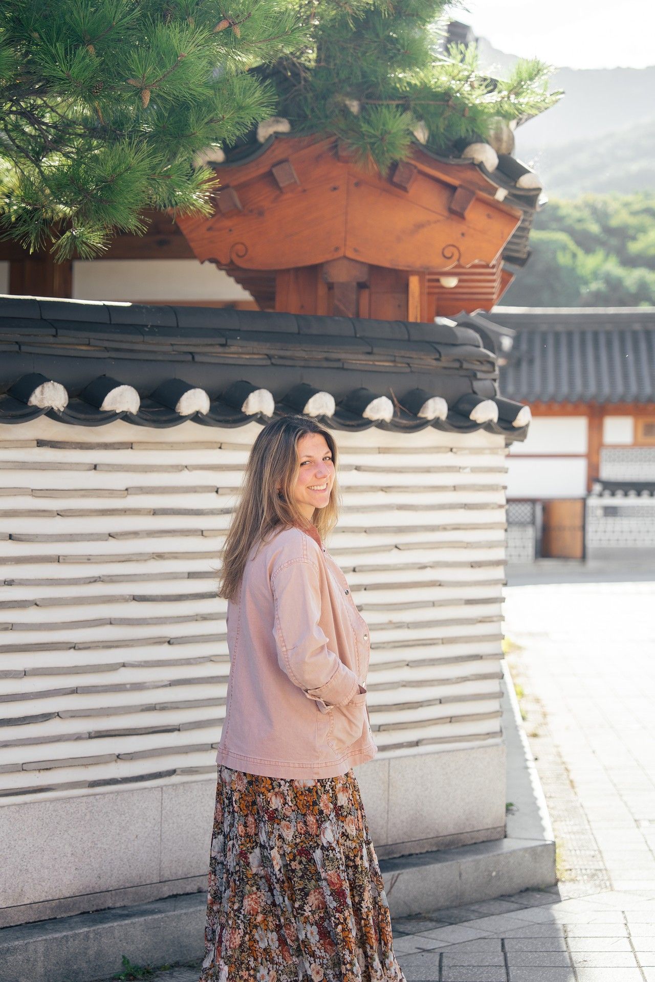 A person in a pink sweater and floral skirt poses against a traditional Korean palace wall with white stone panels.