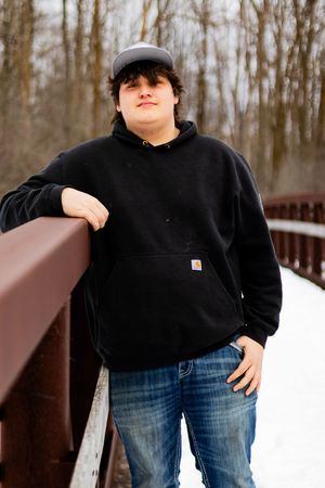 Person in black Carhartt hoodie and jeans posing on wooden bridge in winter.