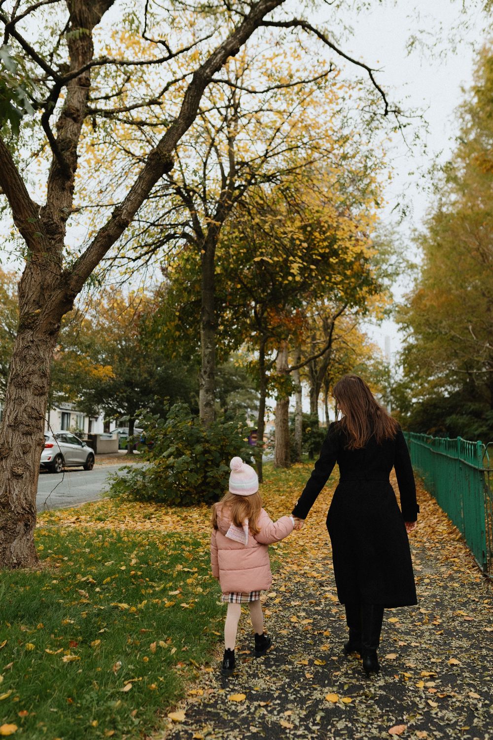 Two people walking along a leaf-covered path on an autumn day with trees displaying golden foliage.