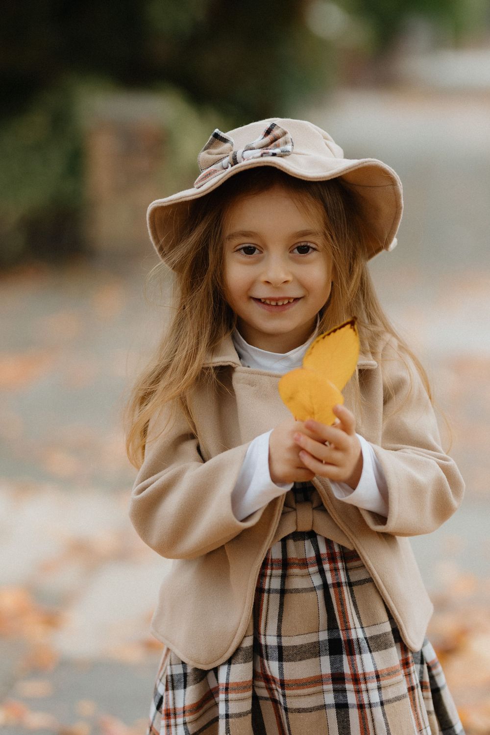 A child in a beige coat and plaid dress plays with autumn leaves in a series of connected outdoor photos.