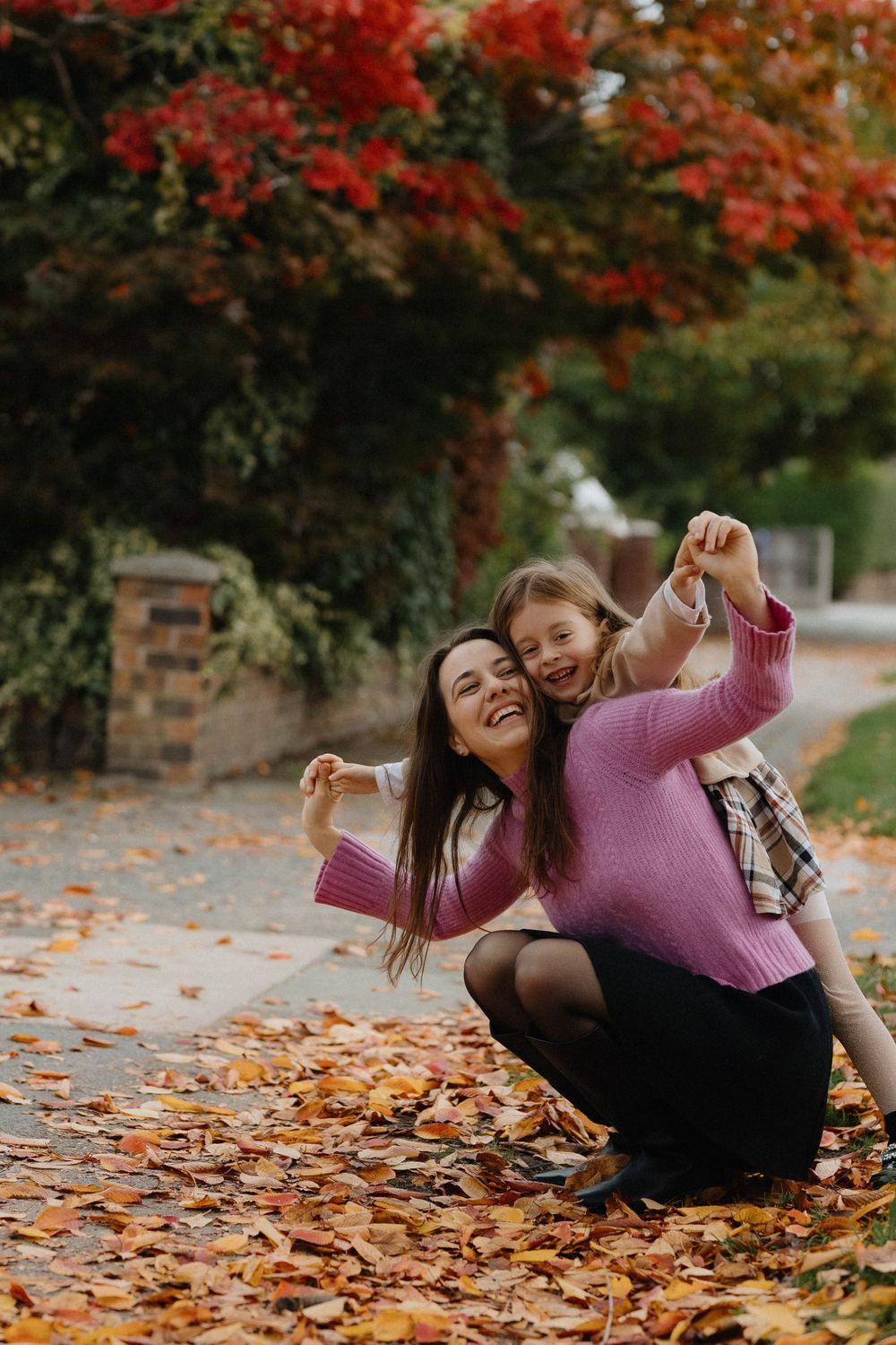 Two people enjoy a playful autumn moment among fallen leaves under trees with vibrant red foliage.