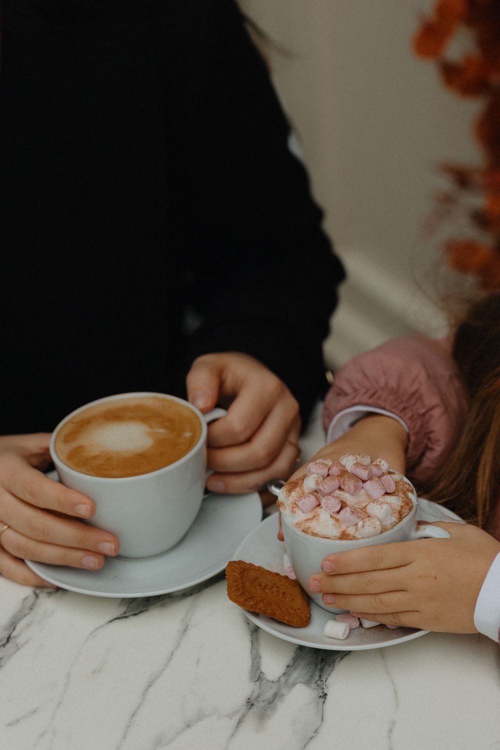 A series of hands holding white coffee cups with latte art and marshmallows on a marble table surface.