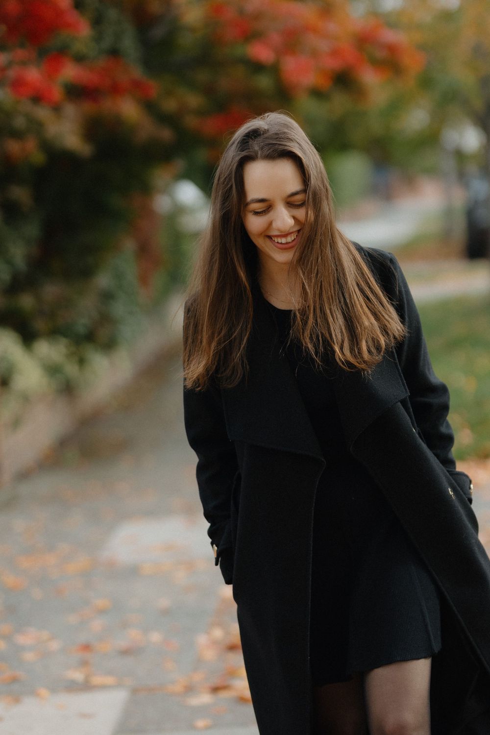 Woman in black coat laughs joyfully while walking through autumn foliage with vibrant red leaves in background.