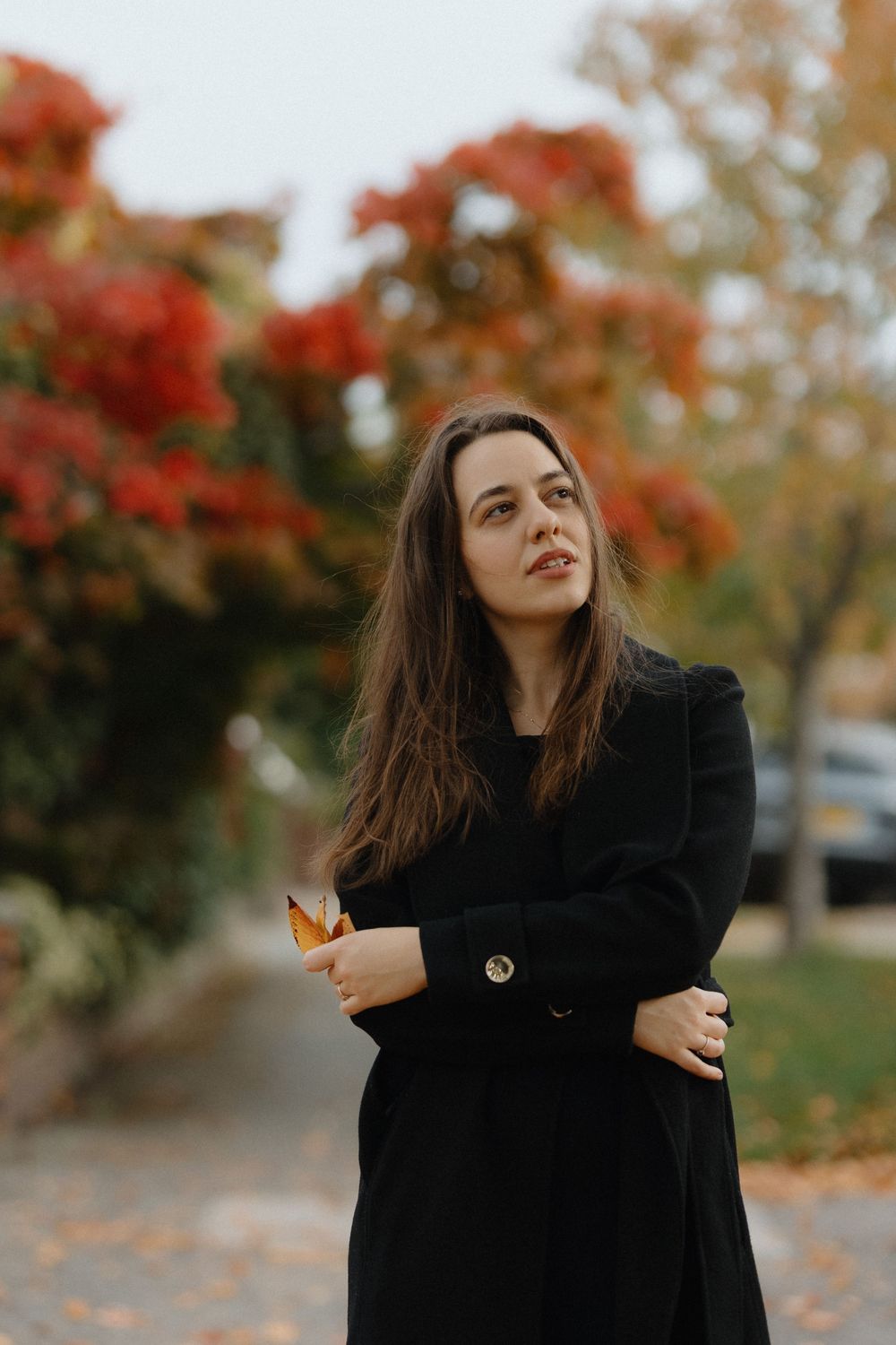 Woman in black coat poses playfully outdoors against a backdrop of fall trees with red autumn leaves.