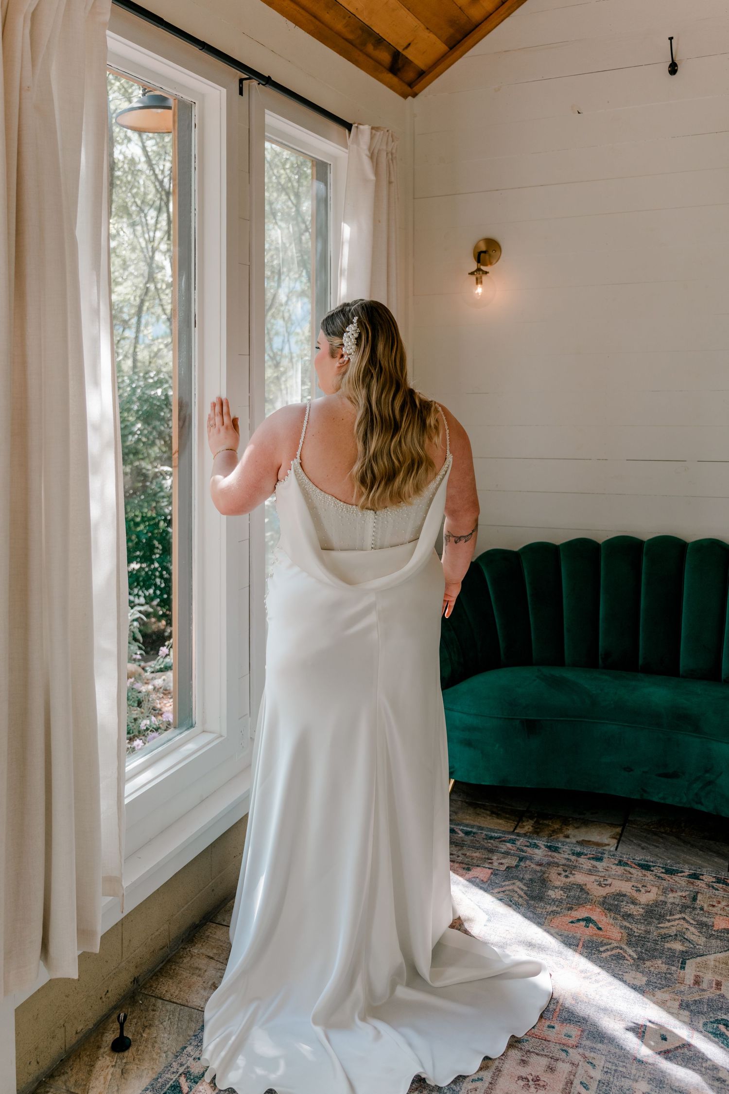 A bride in a flowing white dress stands by large windows in a room with emerald velvet couch and wooden ceiling.