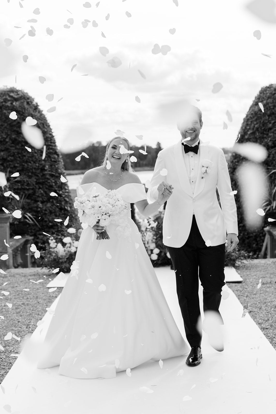 Wedding couple walking outdoors while white flower petals fall around them during a joyful celebration.