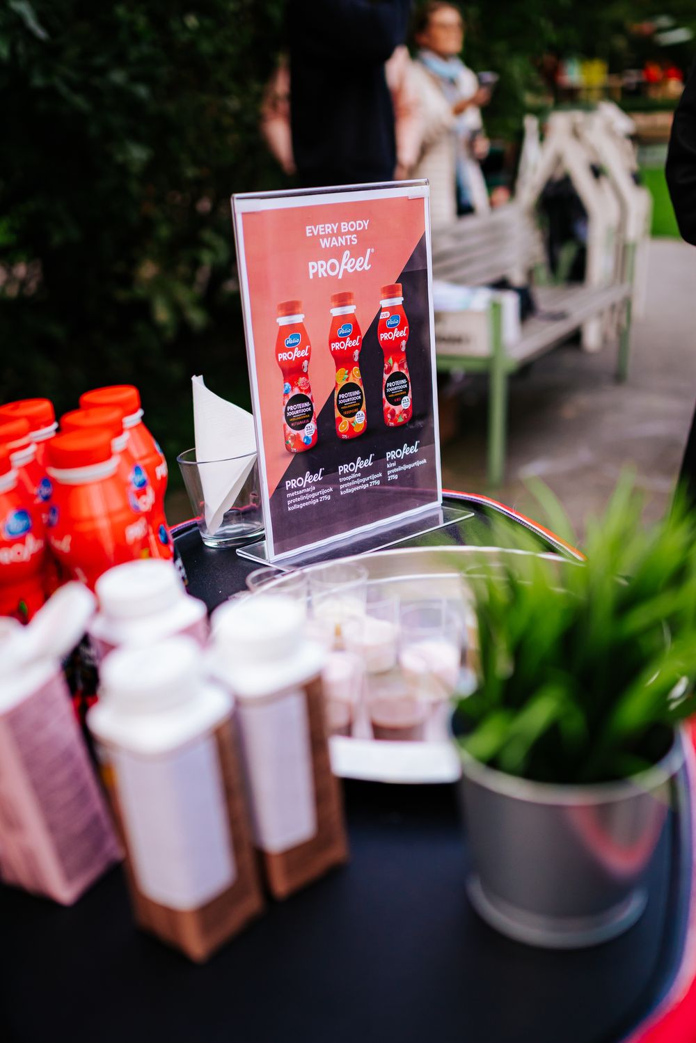 A beverage display with pink bottles and a promotional sign showing different drink options on a dark table with plants nearby.