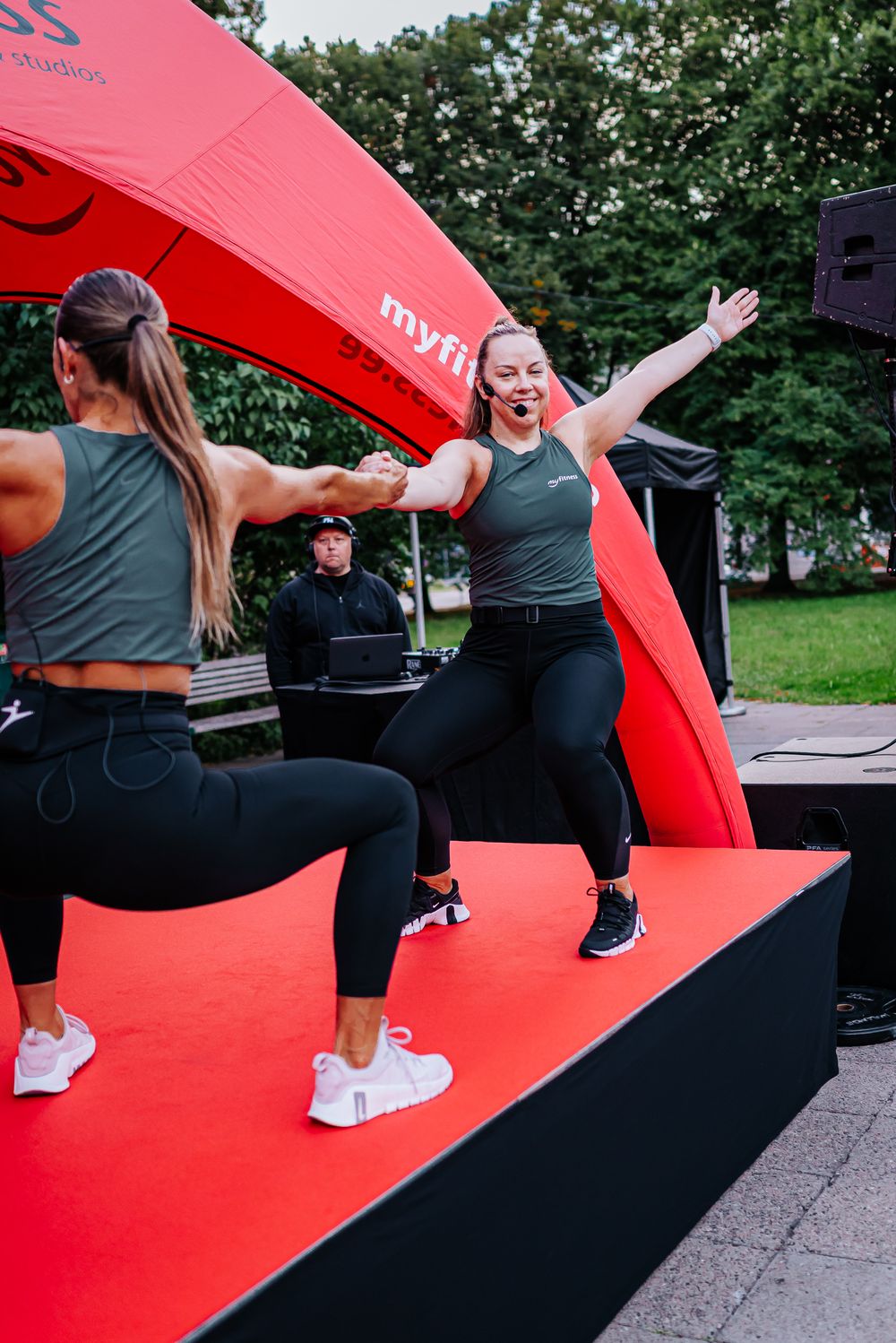 Two fitness instructors lead an energetic workout class on a red stage platform under a bright red canopy outdoors.