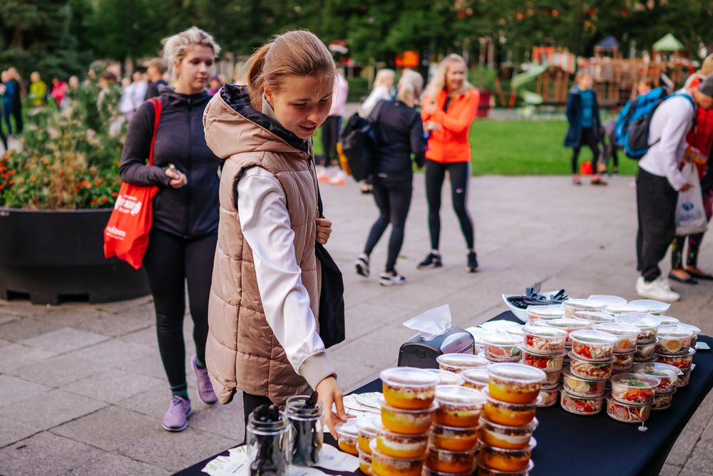People browse food items displayed on a table at an outdoor market or event in a public park.