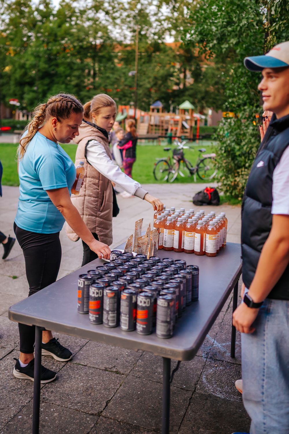 People gathered around a table with drinks and beverages set up at an outdoor event in a park setting.