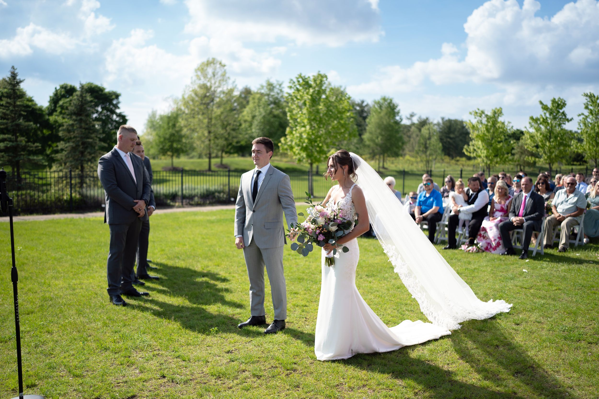 An outdoor wedding ceremony takes place on a sunny day with green grass and trees in the background.