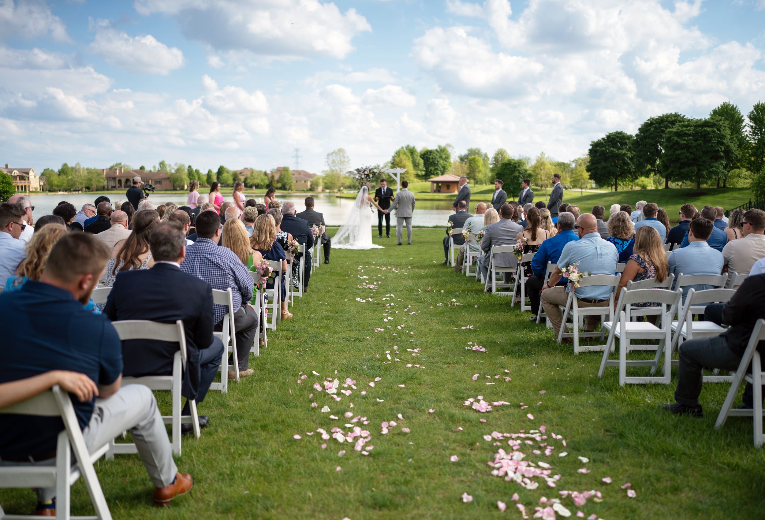 Panoramic view of outdoor wedding ceremony setup with white chairs and flower petals scattered on green grass.