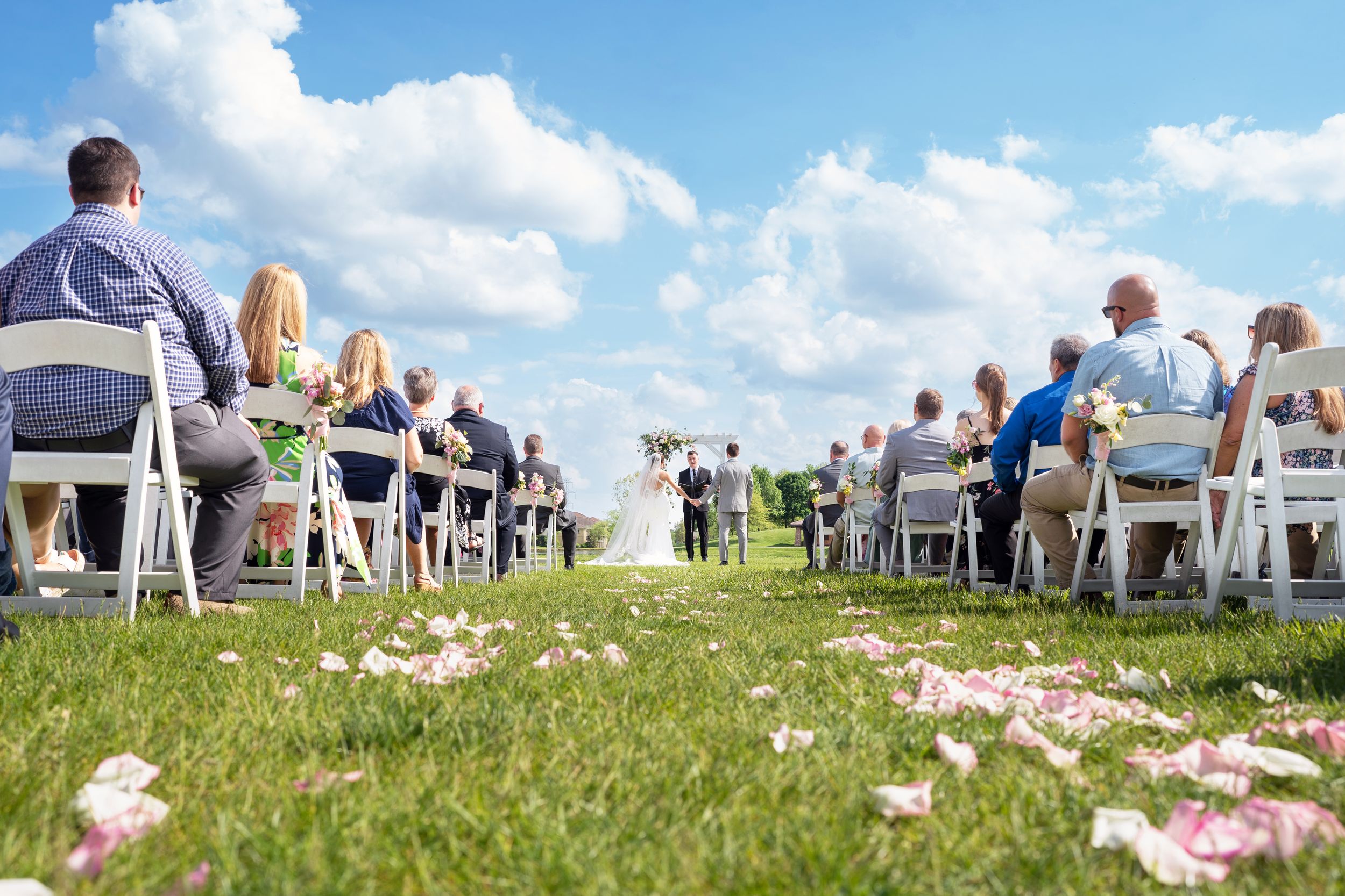Guests seated in rows of white chairs for outdoor wedding ceremony on sunny grassy hillside.