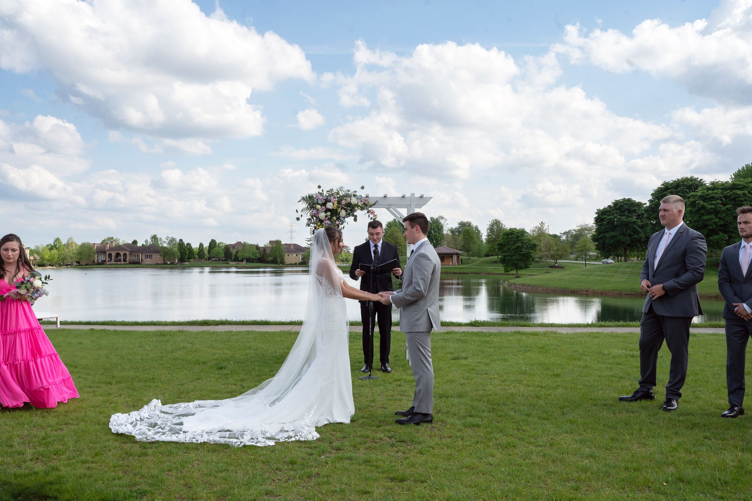 A wedding ceremony takes place on a lush green lawn with white chairs and guests gathered under blue skies.