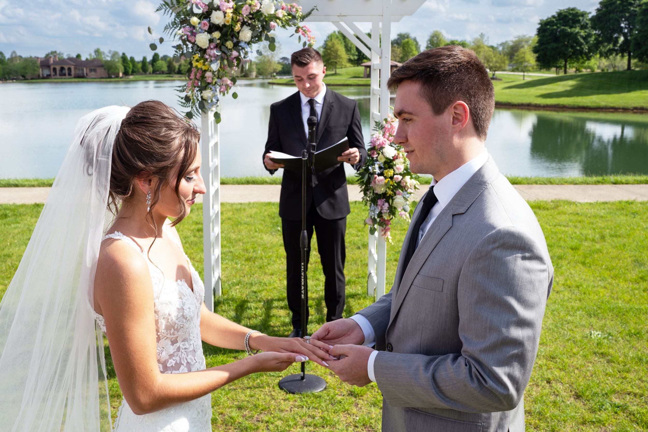 A wedding ceremony takes place outdoors on a sunny day with a lake and trees in the background.