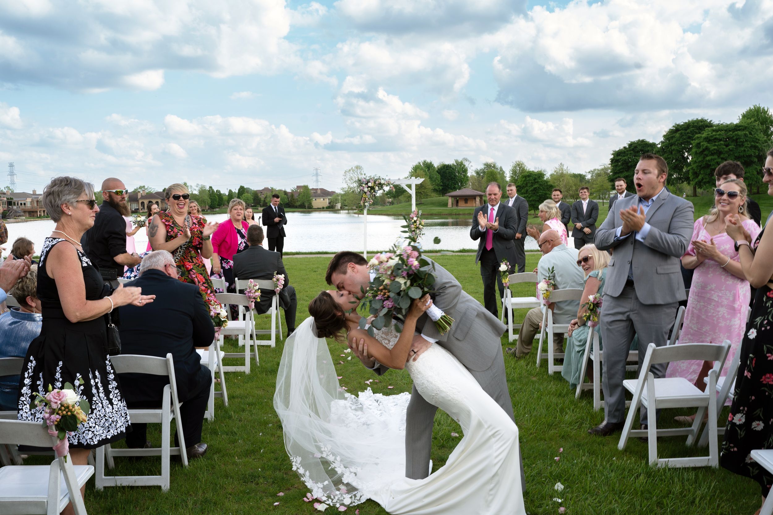 Guests seated in white chairs at an outdoor lakeside wedding ceremony with cloudy skies above.