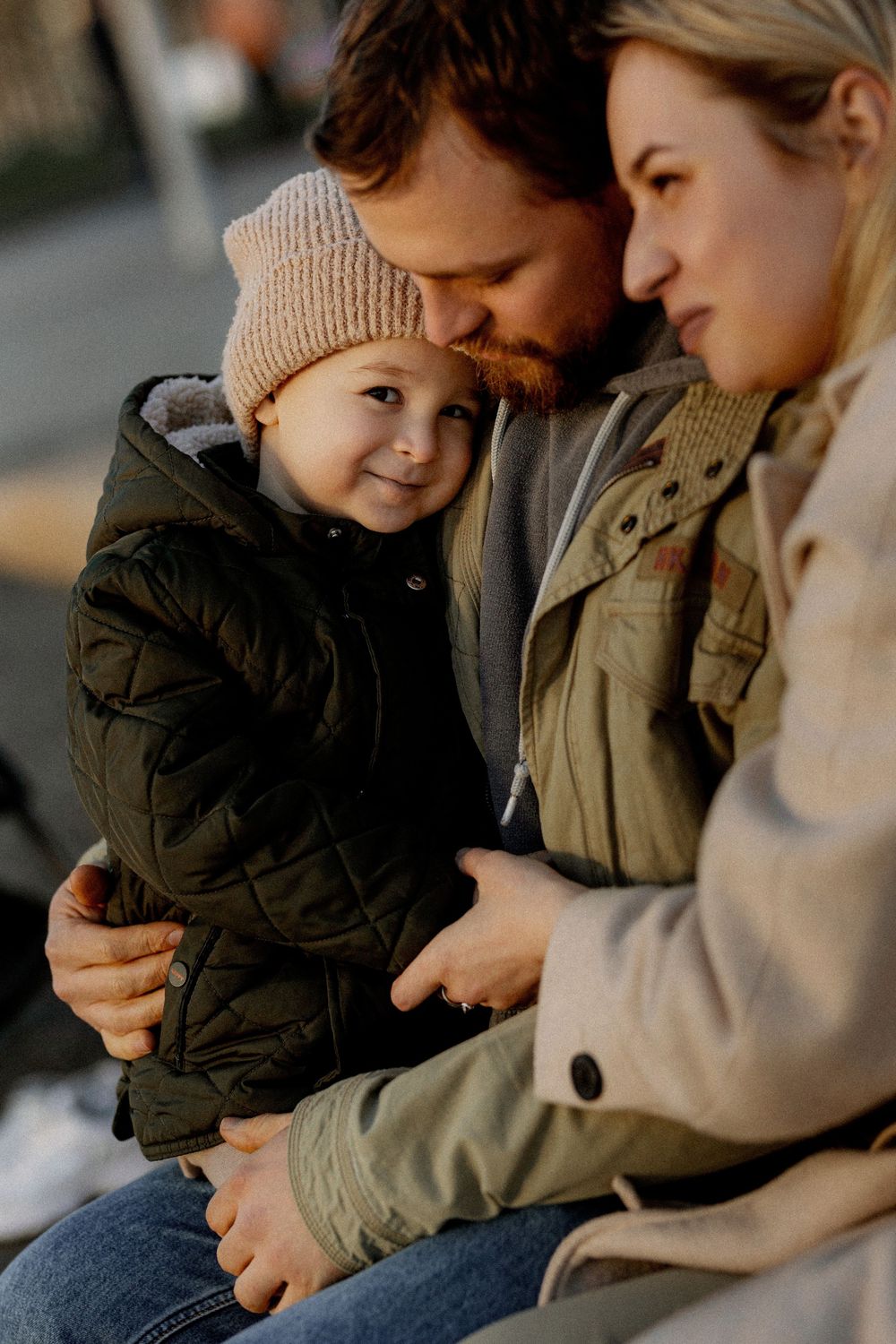A family shares an intimate moment outdoors, with a small child in a knit beanie and quilted jacket being embraced.
