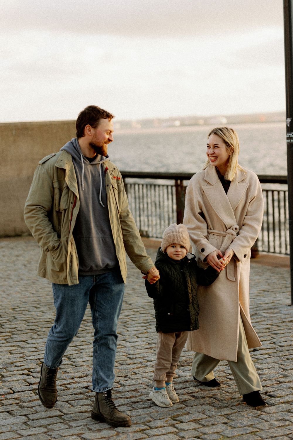 A couple shares a playful moment walking together on cobblestone pavement at sunset by the waterfront.