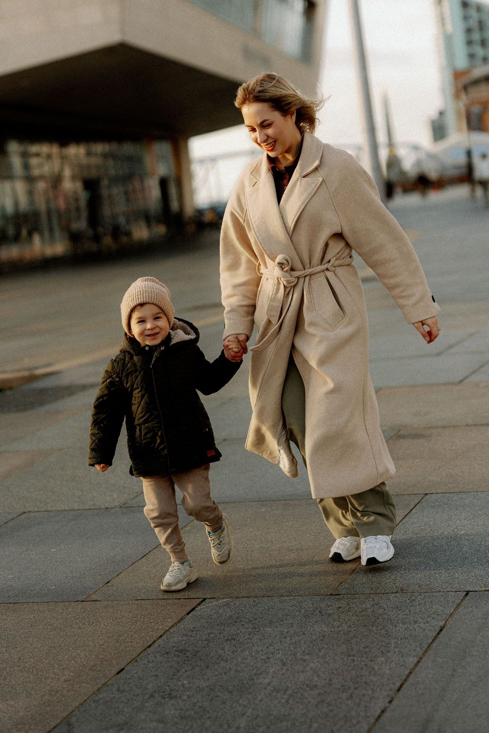 Parent and toddler in winter coats share a playful moment walking together on an urban sidewalk in warm sunlight.