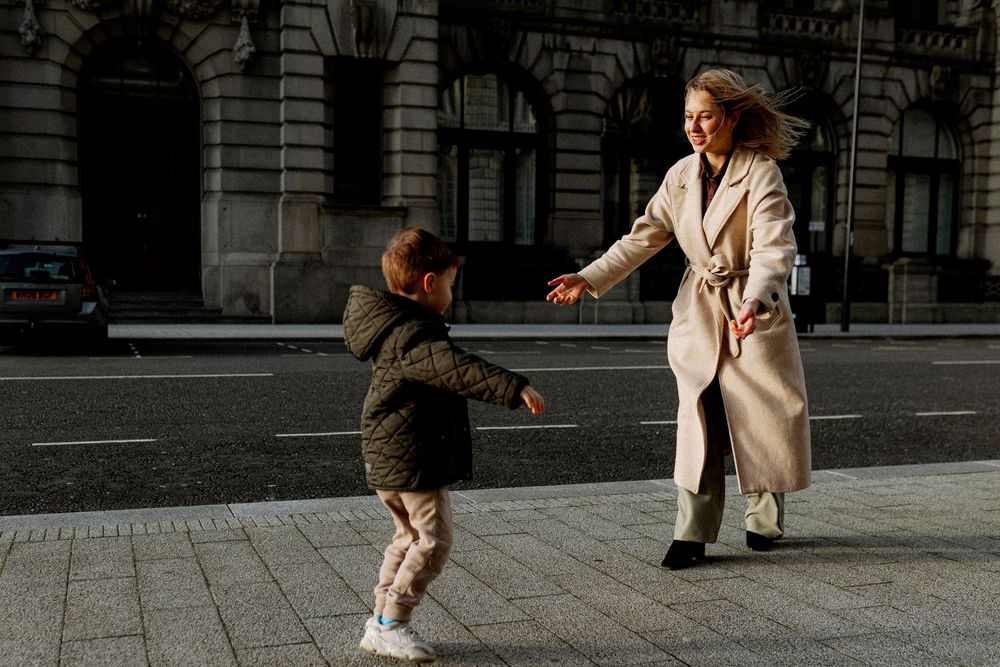 A person in a beige coat and boots performs a dynamic dance move on an urban street.