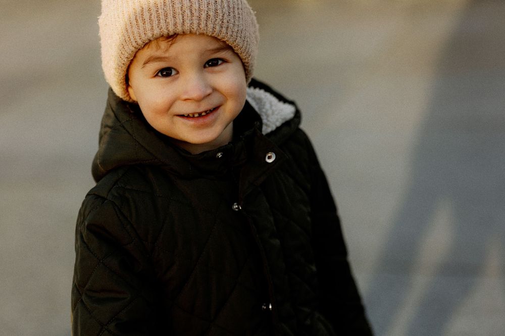 A young child wearing a beige knit beanie and black winter coat smiles warmly on a blurry background.