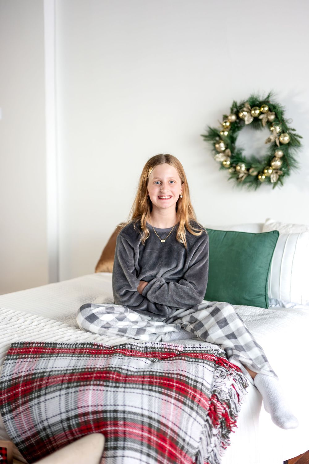 Smiling teenage girl sitting cross-legged on bed with plaid blanket and Christmas wreath backdrop.