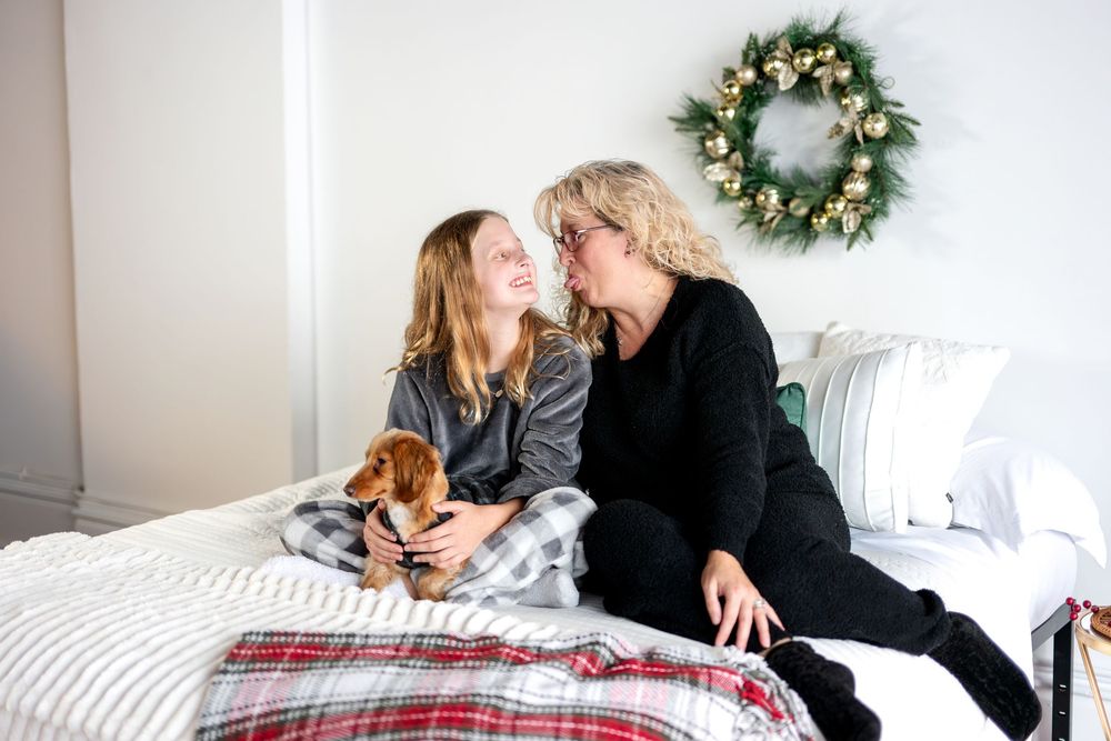 Mother kissing daughters cheek while holding dachshund puppy on bed with Christmas wreath.