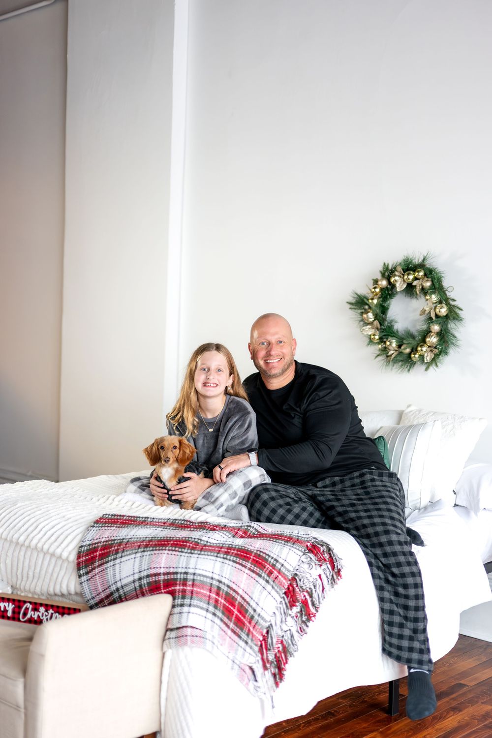 Family in Christmas pajamas sitting on bed with dachshund puppy and holiday wreath backdrop.