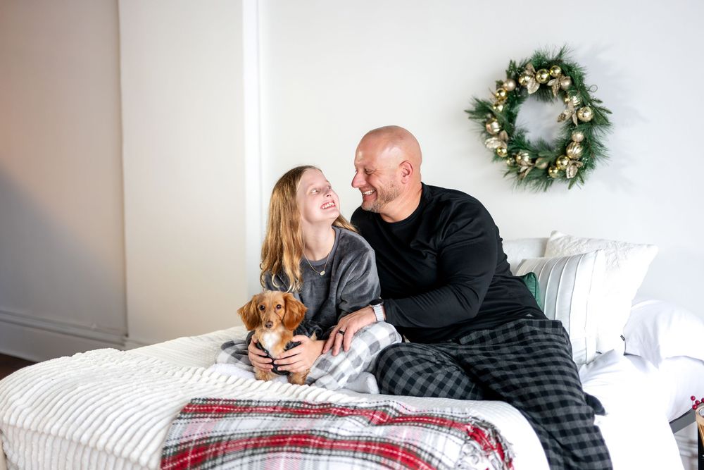 Smiling couple in holiday pajamas sitting on white bed with dachshund and Christmas wreath.