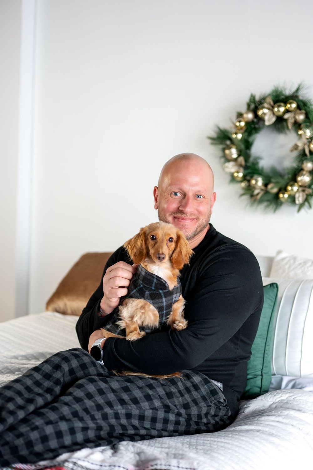 Smiling bald man in black sweater holding dachshund in plaid outfit on bed with Christmas wreath.