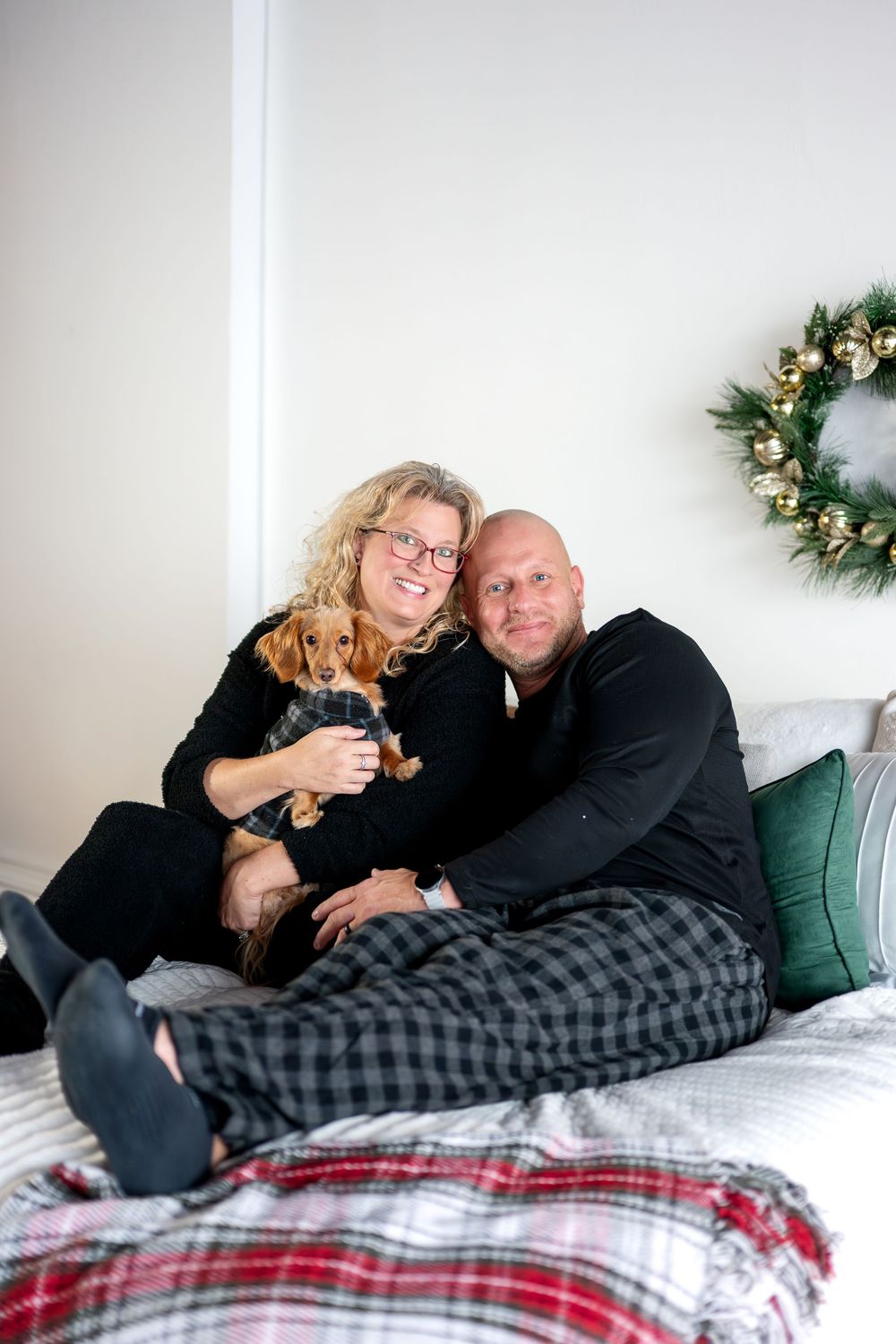 Smiling couple in Christmas pajamas cuddling dachshund puppy on bed with holiday wreath decor.