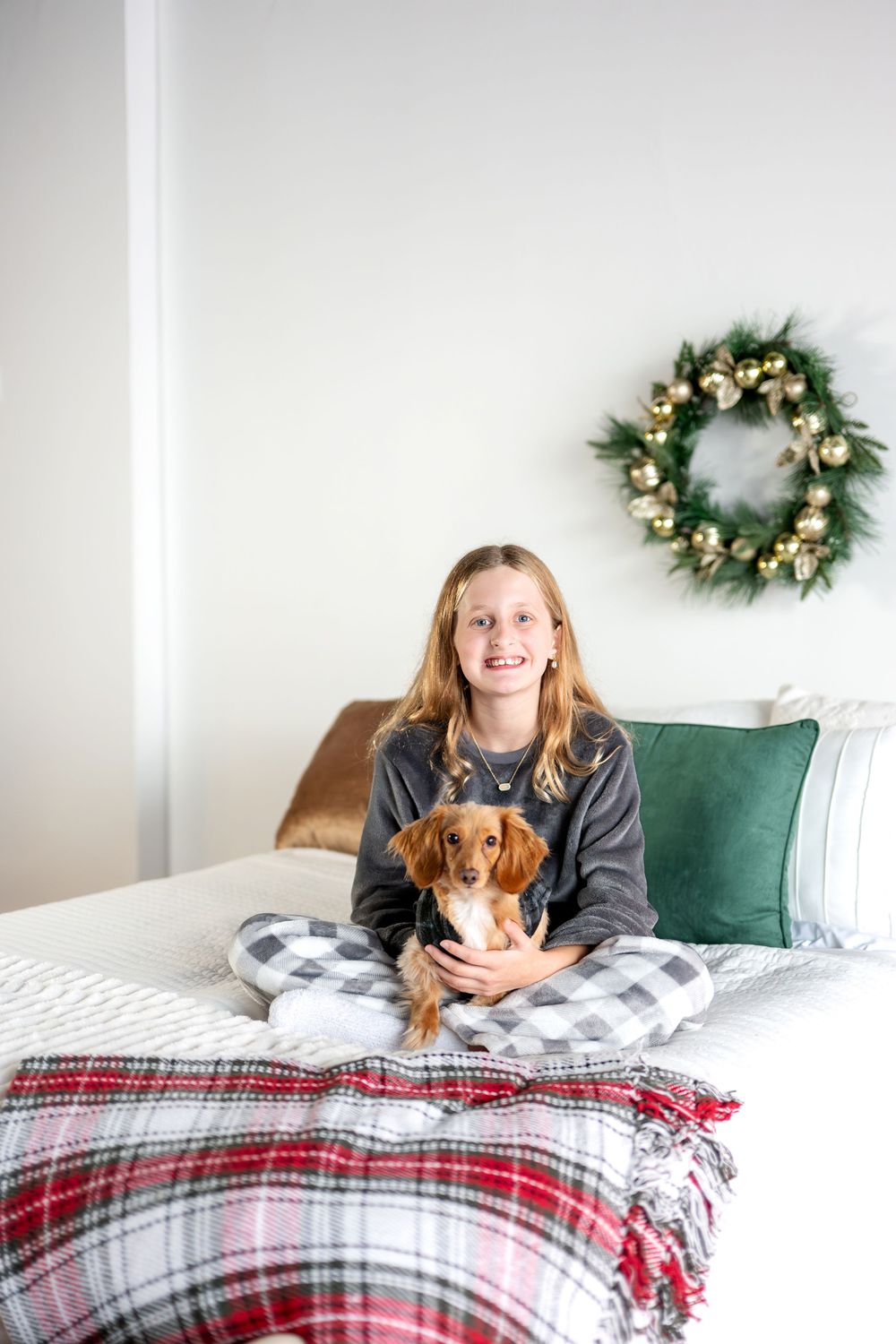 A smiling teenage girl sits cross-legged on a bed holding a small dachshund, with a gold Christmas wreath on the wall.