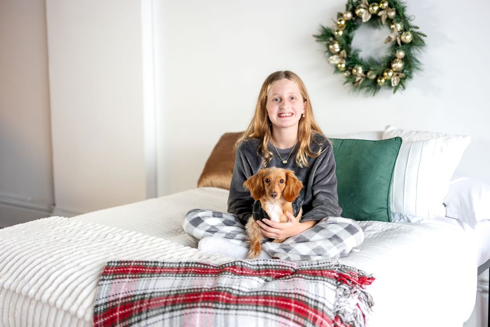 A teenage girl sits on a bed holding a small dachshund dog, with a festive Christmas wreath hanging on the wall behind her.