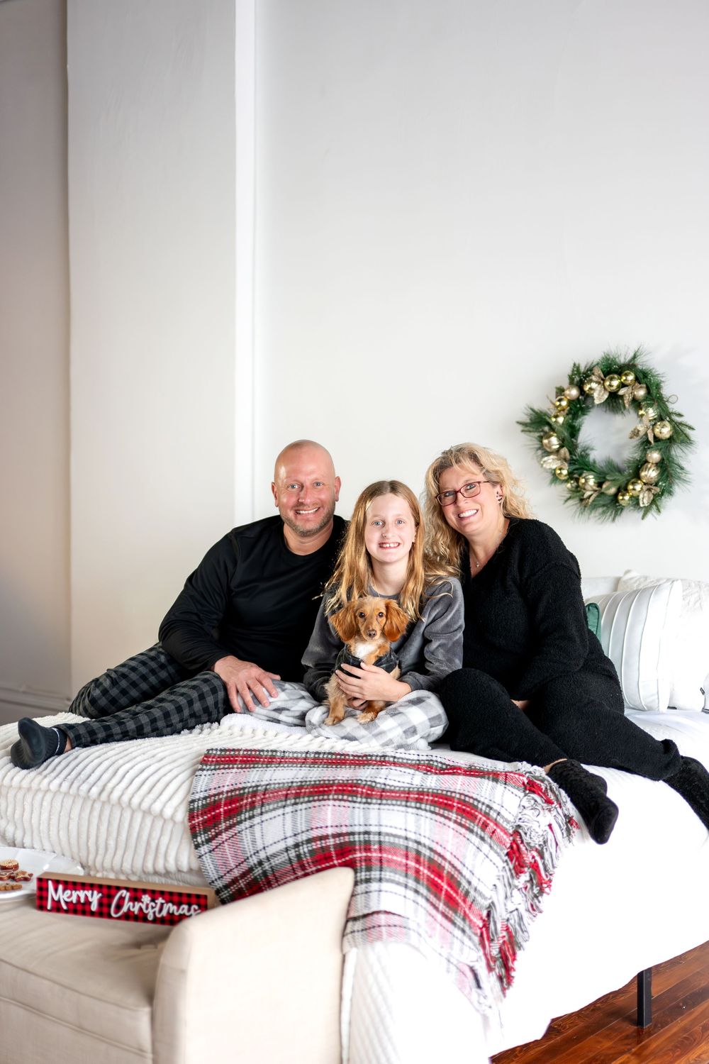 Family of three with dachshund dog posing on bed with Christmas plaid blanket and holiday wreath.
