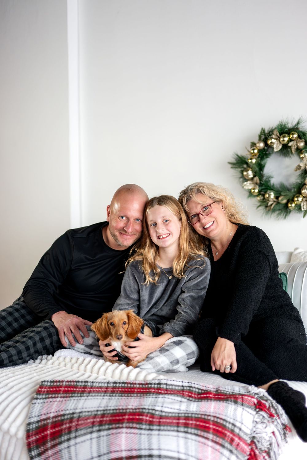 Cheerful family of three with dachshund dog posing on bed with festive Christmas wreath.