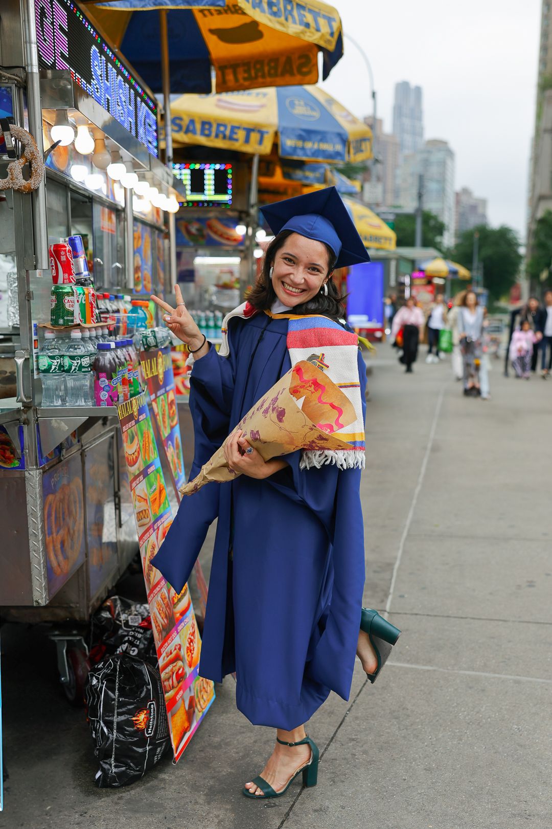 Graduate in blue cap and gown poses with flowers by street vendor cart in city with skyscrapers in background.