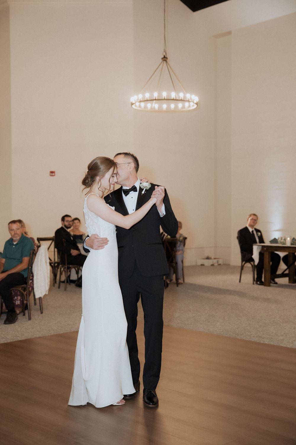 A couple shares their first dance in an elegant reception hall with a modern circular chandelier overhead.