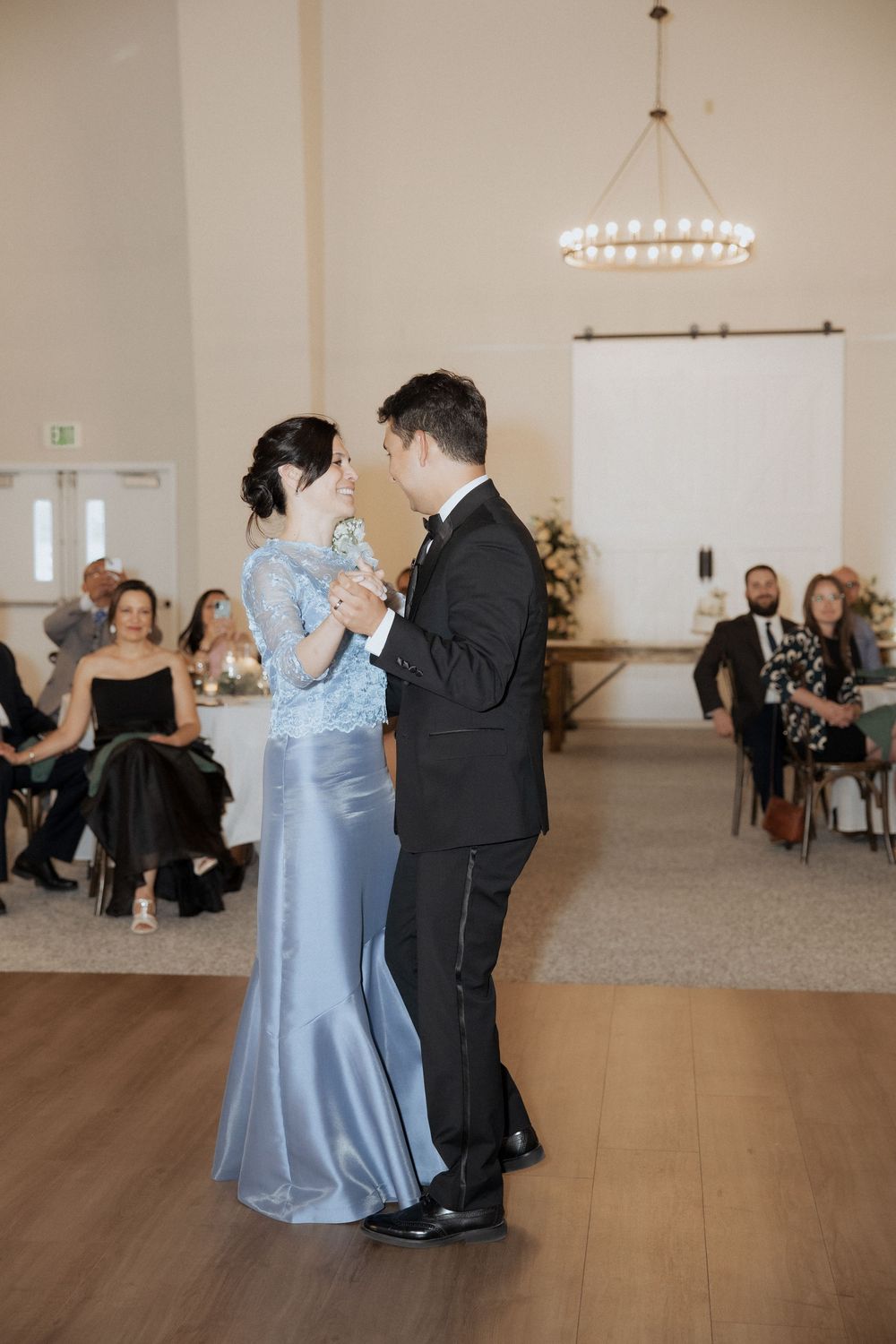 Couple dancing together in a light-filled reception hall with guests seated in the background.