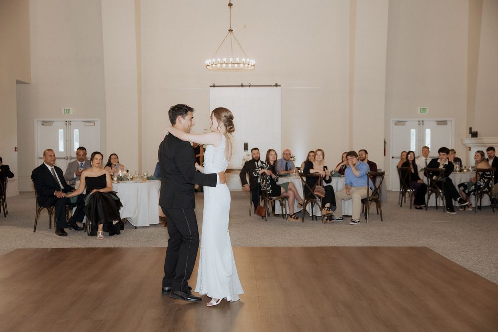 Newlywed couple shares their first dance in an elegant white ballroom surrounded by seated wedding guests.