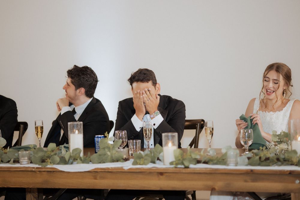 People seated at a long wooden table decorated with greenery and floral arrangements for a wedding reception.