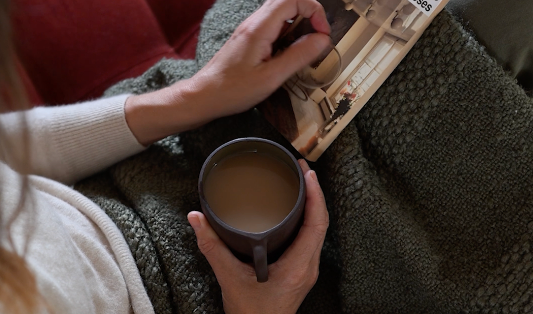 Cosy setting with woman browsing a magazine and holding a cup of coffee on a beautiful velvet sofa with a green blanket