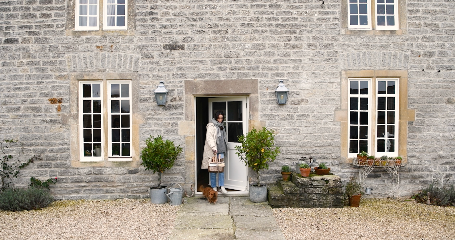 Woman leaves her rustic stone cottage with white-framed windows and potted plants flanking the entrance on a gravel path.