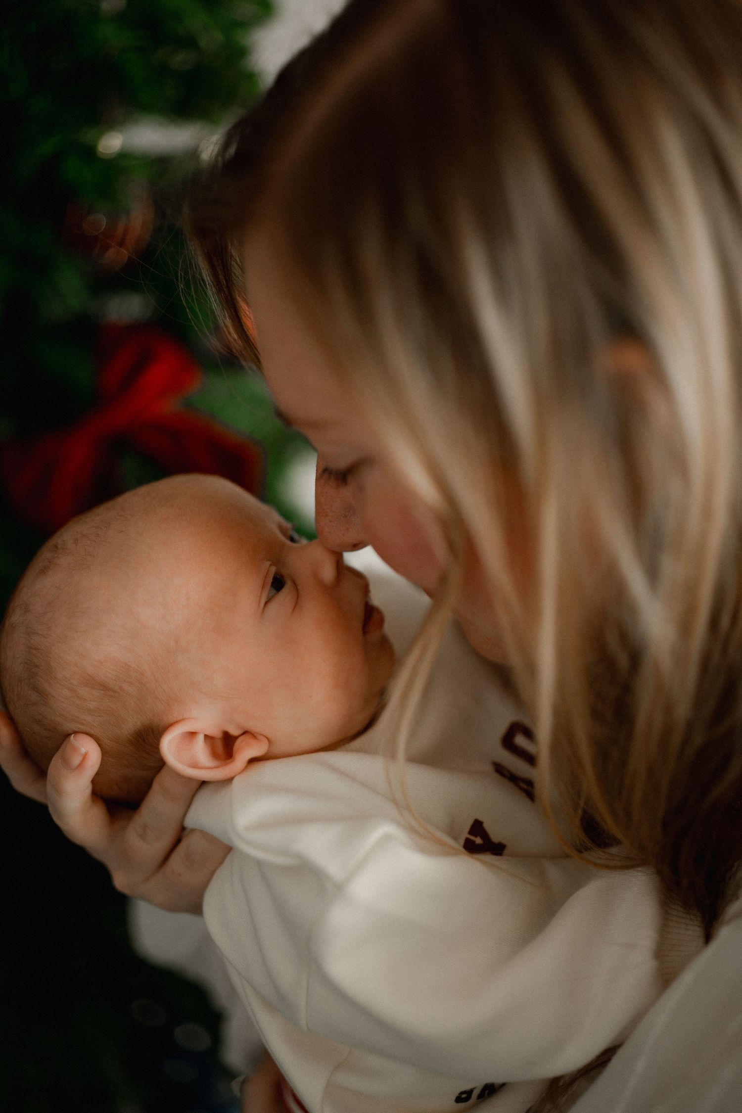 A tender moment as a blonde mother touches noses with her newborn baby near a Christmas tree.