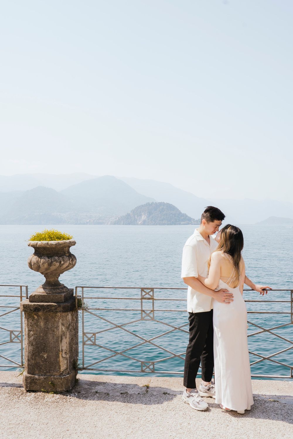 A romantic couple shares a tender moment by a lakeside stone railing with mountains in the background.