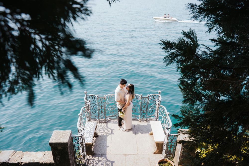 A romantic couple shares a moment on a white dock overlooking turquoise waters and evergreen trees.