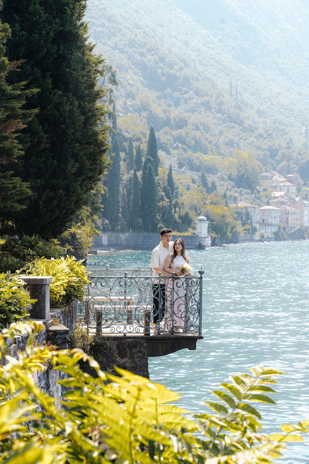 A couple stands on an ornate balcony overlooking Lake Como with cypress trees and mountains in the background.